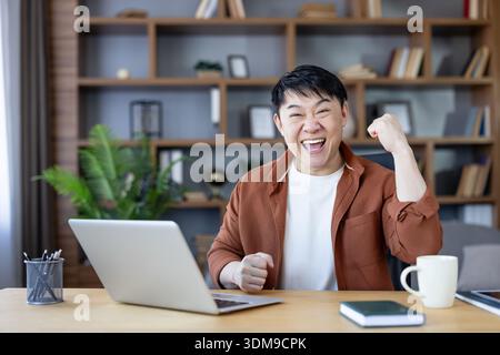 Heureux homme asiatique avec un sourire authentique et levé le poing avec enthousiasme célébrant le succès des affaires tout en travaillant à la maison, en atteignant un objectif et en se sentant de la joie à son bureau Banque D'Images