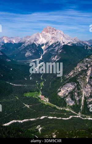 Vue aérienne d'une route sinueuse à travers une vallée forestière menant au pic massif de Croda Rossa au loin, vu de Monte Piana. Banque D'Images