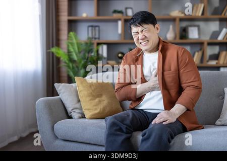 Homme asiatique souffrant d'une crise cardiaque soudaine ou d'une angine de poitrine alors qu'il était assis sur un canapé de salon, serrant sa poitrine avec une expression d'inconfort intense et d'urgence médicale Banque D'Images