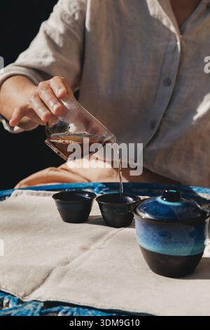 Une photo en gros plan capture une cérémonie sereine du thé alors qu'une main douce verse un riche thé ambré d'un récipient en verre dans deux élégantes tasses en céramique. Banque D'Images