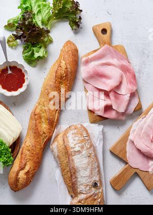 Les ingrédients frais sont disposés sur une table. Une baguette, des tranches de jambon, du fromage mozzarella, de la laitue verte et de la sauce tomate sont prêts. La scène montre un Banque D'Images