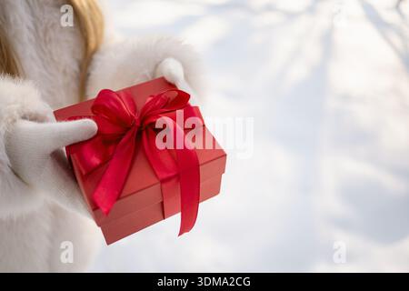 Gros plan de la boîte-cadeau rouge dans les mains de la femme portant des gants d'hiver, vacances et concept de cadeau saisonnier à l'extérieur. Banque D'Images
