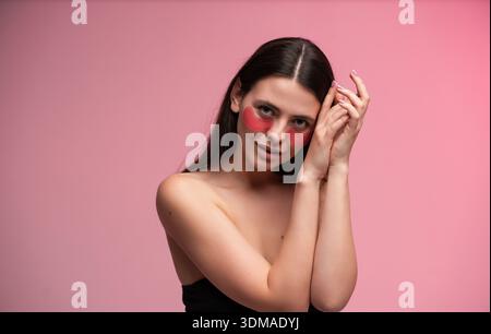 Jeune femme avec de longs cheveux foncés levant la main et portant rose sous des patchs oculaires en studio sur fond rose doux, concept de soins de la peau Banque D'Images