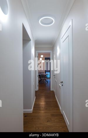 Un couloir étroit avec des murs blancs et des portes lambrissées mène à une salle à manger. Les planchers en bois chauds contrastent avec un anneau LED froid et des appliques minimales, suggérant Banque D'Images