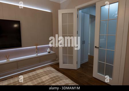 Chambre beige douce dans un appartement montre des accents LED, armoires encastrées, TV murale, couette à carreaux, portes françaises givrées aux placards de couloir, plancher de bois chaud, Banque D'Images