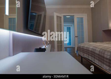 Vue au rez-de-chaussée d'une chambre beige avec lit à carreaux, armoires grises et portes doubles françaises. Une télévision murale et une console nervurée éclairée par LED avec diffuseurs appe Banque D'Images
