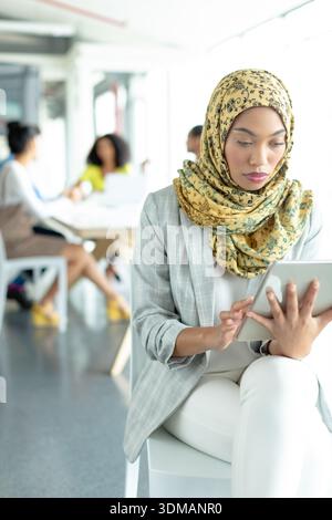 Femme en jaune-hijab, blazer gris, assise sur une chaise blanche à l'aide d'une tablette au bureau avec un travail d'équipe. Collègues, travail d'équipe, collaboration, productivité Banque D'Images