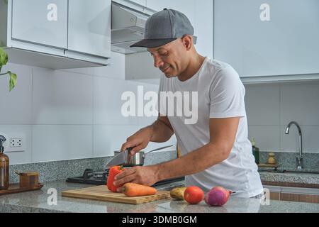 Homme tranchant des légumes sur une planche à découper, préparant un repas sain dans une cuisine moderne Banque D'Images