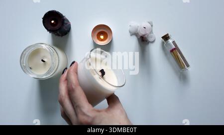 Entretien de la bougie : vue de dessus de la main d'une femme tenant une bougie parfumée au soja naturel dans un bocal en verre. A proximité se trouvent d'autres bougies colorées et une bouteille en verre avec Banque D'Images