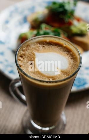Une tasse de café avec un coeur dessiné sur le dessus est assise sur une table à côté d'une assiette de nourriture. Le café est brun et mousseux, et le cœur est blanc et légèrement Banque D'Images