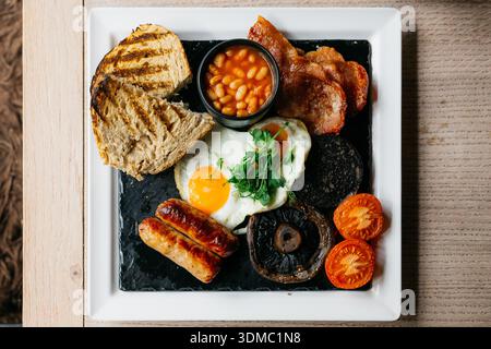 Petit-déjeuner anglais complet traditionnel avec œufs frits saucisses bacon haricots pain grillé et légumes grillés, présentés sur une assiette en ardoise pour les menus du café, Banque D'Images