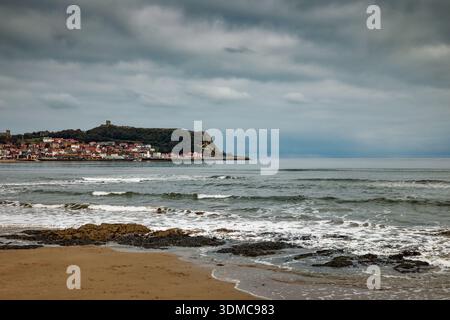 De douces vagues se jettent sur une plage par une journée nuageuse avec la station balnéaire de Scarborough au loin et le château sur le promontoire, North Yorkshire. Banque D'Images