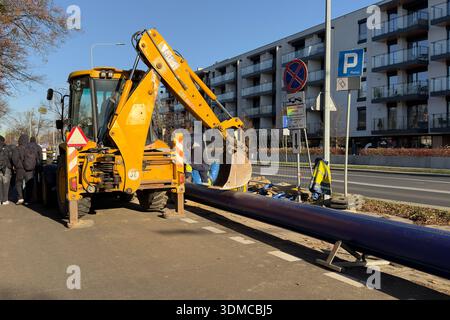 Poznan, Pologne - 18 novembre 2025 : une chargeuse-pelleteuse jaune vif avec son godet d'excavation abaissé près d'un long tuyau bleu, prête pour la déve urbaine Banque D'Images