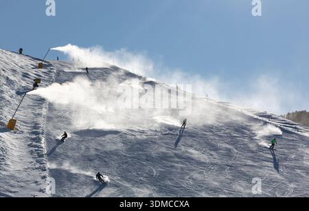 Skieurs descendant une pente enneigée raide avec de la neige artificielle provenant de machines à neige lors d'une journée d'hiver ensoleillée. Banque D'Images