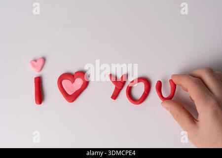 Une représentation ludique et créative du message « je t'aime » faite d'argile rouge sur une surface blanche, avec un cœur rose et un soupçon d'implication humaine. Banque D'Images