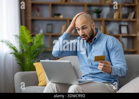 Jeune homme tenant sa carte de crédit en or et plaçant une main sur sa tête, paraissant choqué et stressé tout en éprouvant un problème financier ou une fraude en ligne en utilisant un ordinateur portable à la maison Banque D'Images
