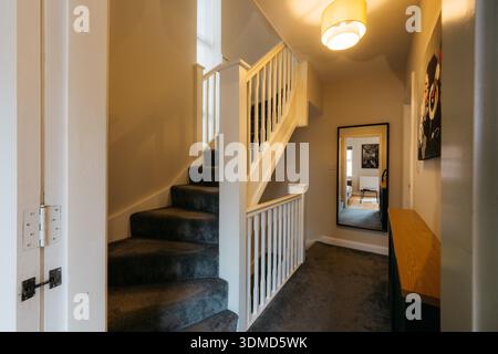 Un couloir avec un escalier et un miroir. L'escalier est en bois et recouvert de tapis. Le couloir est bien éclairé et a une ambiance moderne Banque D'Images