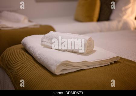 Une serviette blanche est couchée sur un lit avec un oreiller brun. La serviette est enroulée et placée sur le dessus du lit Banque D'Images