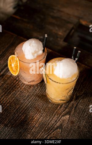Deux verres de boissons mélangées avec glace et garniture de fruits sur une table en bois. Les boissons sont roses et orange Banque D'Images