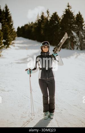 Une skieuse heureuse se tient fièrement sur le sentier enneigé, tenant ses skis, prête pour l'aventure dans un paysage hivernal. Banque D'Images