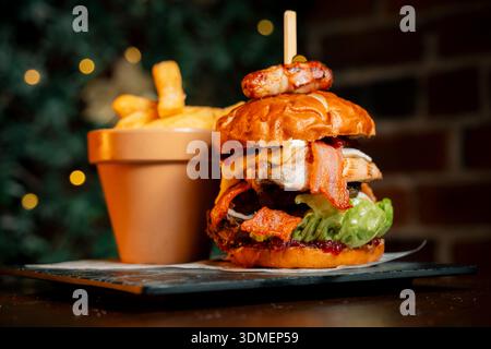 Un grand hamburger au bacon et à la laitue se trouve sur une assiette à côté d'une petite casserole. Le hamburger est surmonté d'un petit pain et d'une lance de cornichon. Concept d'indulgence a Banque D'Images