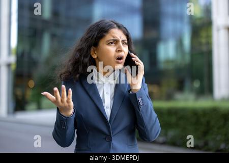 Femme d'affaires indienne à l'extérieur cherchant inquiet et frustré lors d'un appel téléphonique urgent, réagissant aux mauvaises nouvelles tout en gérant le stress et les défis dans un lieu de travail urbain moderne Banque D'Images