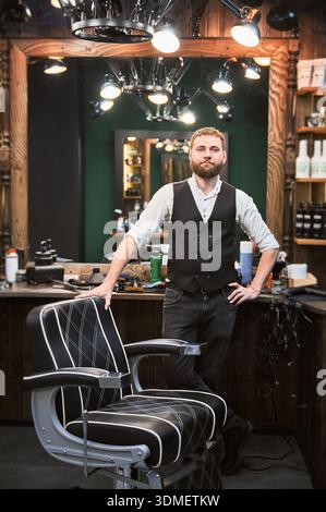 Coiffeur confiant dans une tenue élégante attendant le prochain client. Barbier masculin penchant sa main sur la chaise et regardant la caméra. Travailler dans un barbier moderne. Banque D'Images