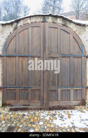 De grandes portes en bois vieilli, situées dans un mur de pierre courbé avec des charnières en fer et un sol poussiéreux de neige, évoquant le charme rustique et un sens de la courtoisie historique Banque D'Images