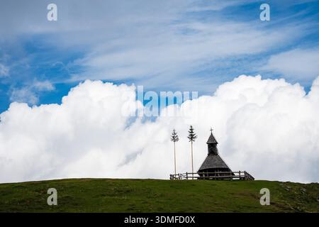 Une petite chapelle traditionnelle en bois avec une croix se trouve sur une crête herbeuse verte sous un ciel bleu vif avec de grands nuages moelleux blancs en Slovénie Banque D'Images