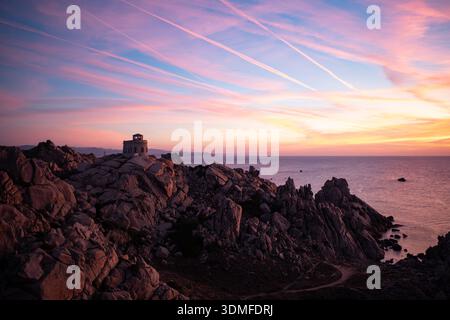 Côte rocheuse au coucher du soleil avec un petit belvédère en pierre surplombant la mer calme, sous des cieux roses et bleus striés de nuages doux. Banque D'Images