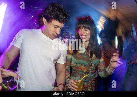 Jeune homme et femme profitant d'une soirée animée, célébrant avec des amis tout en buvant des cocktails dans un environnement de club dynamique avec des lumières énergiques Banque D'Images