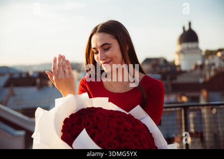 Une femme est heureuse en regardant son anneau tout en tenant un grand bouquet de roses, avec des bâtiments de la ville en arrière-plan. Banque D'Images