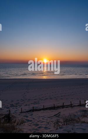 Le soleil se trouve bas sur l'horizon de la mer Baltique en Lituanie, jetant un chemin doré sur les vagues douces, les dunes ondulées, les herbes, les empreintes de pas et un bois flotté Banque D'Images