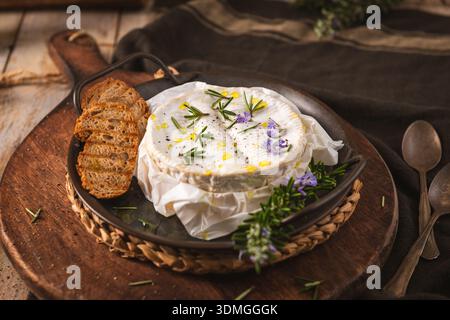 Un fromage camembert gourmand recouvert de brins de romarin frais, de fleurs comestibles et d'huile d'olive. Servi dans une poêle en fonte rustique avec baguet croustillant Banque D'Images