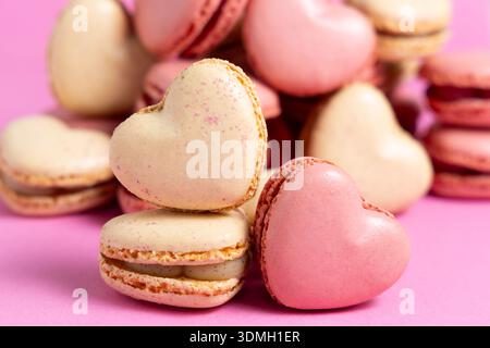 Un groupe de macarons en forme de cœur de différentes couleurs est disposé sur un fond rose vif. Banque D'Images