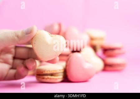 Une main tient un macaron en forme de cœur tandis que d’autres macarons reposent sur une surface rose. Banque D'Images