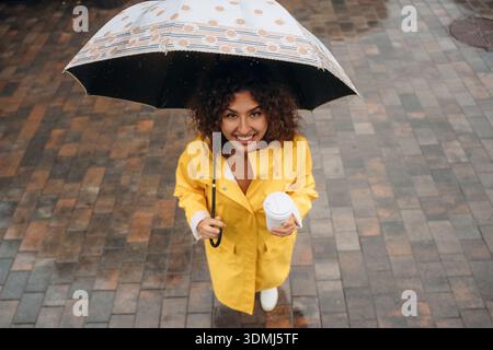 Vue de dessus de la femme qui est debout avec un parapluie et une tasse de café dans une ville pluvieuse, sur les pavés. Banque D'Images