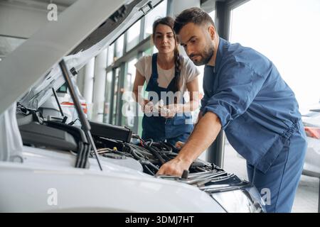 Vue de côté, le capot est ouvert. Les employés de l'atelier de réparation automobile travaillent ensemble. Homme et femme. Banque D'Images