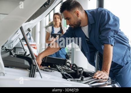 Vue de côté, le capot est ouvert. Les employés de l'atelier de réparation automobile travaillent ensemble. Homme et femme. Banque D'Images