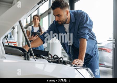 Vue de côté, le capot est ouvert. Les employés de l'atelier de réparation automobile travaillent ensemble. Homme et femme. Banque D'Images