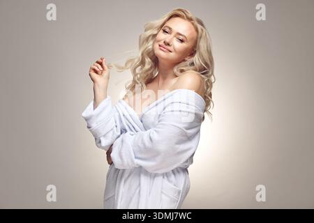 La personne féminine senior dans le peignoir spa a une peau saine parfaitement propre. Belle femme blonde mature portant dans la maison sur gris isolé Banque D'Images
