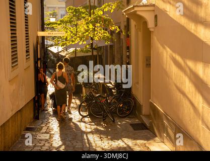 Ljubljana, Slovénie - 10 août 2025 : les piétons traversent une ruelle étroite et ensoleillée à Ljubljana où la lumière de l'heure d'or illumine les pavés Banque D'Images