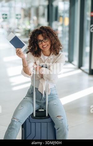Montrant le document, assis sur la valise. Belle femme est dans le hall de l'aéroport. Banque D'Images