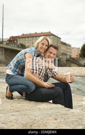 Heureux couple embrassant tout en étant assis au bord de la rivière avec du café Banque D'Images