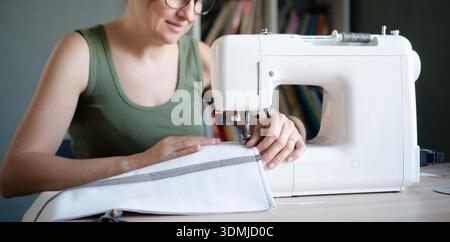 Gros plan d'une couturière joyeuse souriante tout en travaillant à sa machine à coudre dans le studio, aimant son métier. Banque D'Images