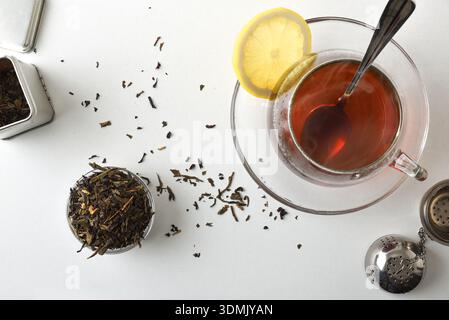 Tasse en verre fraîchement infusée sur une assiette sur un banc de cuisine blanc avec infuseur et récipients pleins d'herbes de thé séchées. Vue de dessus. Banque D'Images