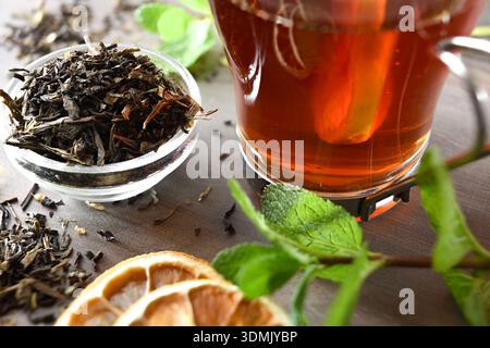 Détail du bol plein de feuilles de thé séchées et infusion de thé noir dans une tasse en verre avec des tranches de citron et de menthe sur une table en bois. Vue surélevée. Banque D'Images