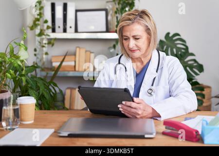 Médecin féminin expérimenté dans le manteau blanc en utilisant la tablette dans le cadre moderne de cabinet médical pendant la consultation du patient Banque D'Images