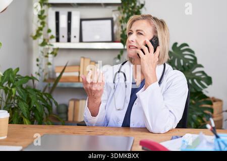 Médecin senior au téléphone dans le bureau, conversation médicale, professionnel attentionné en manteau blanc Banque D'Images