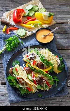 Tacos en boulettes de viande avec chou croquant, tranches de concombre, poivron rouge et jaune, laitue sur une assiette noire sur une table en bois rustique avec des ingrédients, Banque D'Images